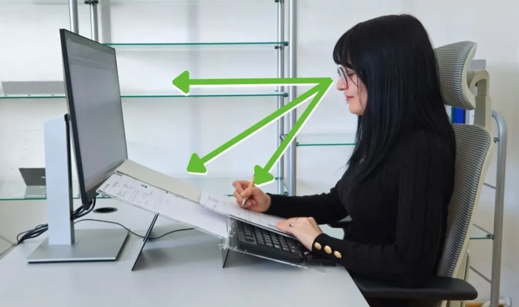 Oseba sedi za delovno mizo v pravilni ergonomski drži, z vzravnano hrbtenico in podprtim hrbtom, medtem ko uporablja računalnik in piše na dokument. Zelene puščice prikazujejo ustrezne smeri pogleda med monitorjem, dokumentom in delovno površino, kar ponazarja ergonomično ureditev delovnega mesta.